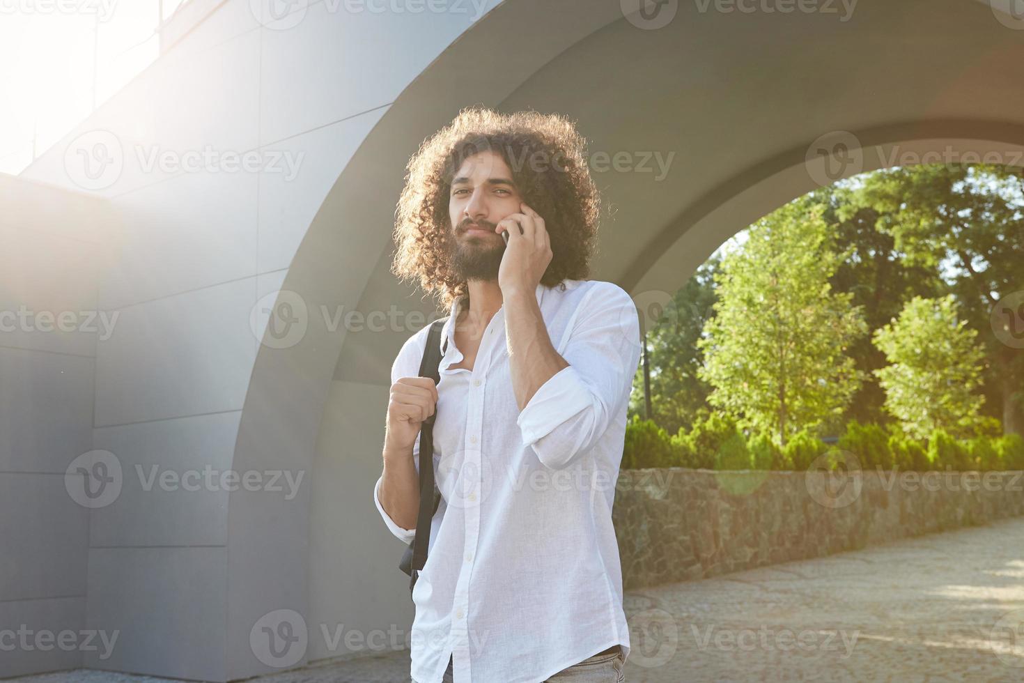 Pretty young curly man with beard walking through city green park on warm sunny day, giving call with his smart phone, wearing white shirt and black backpack photo