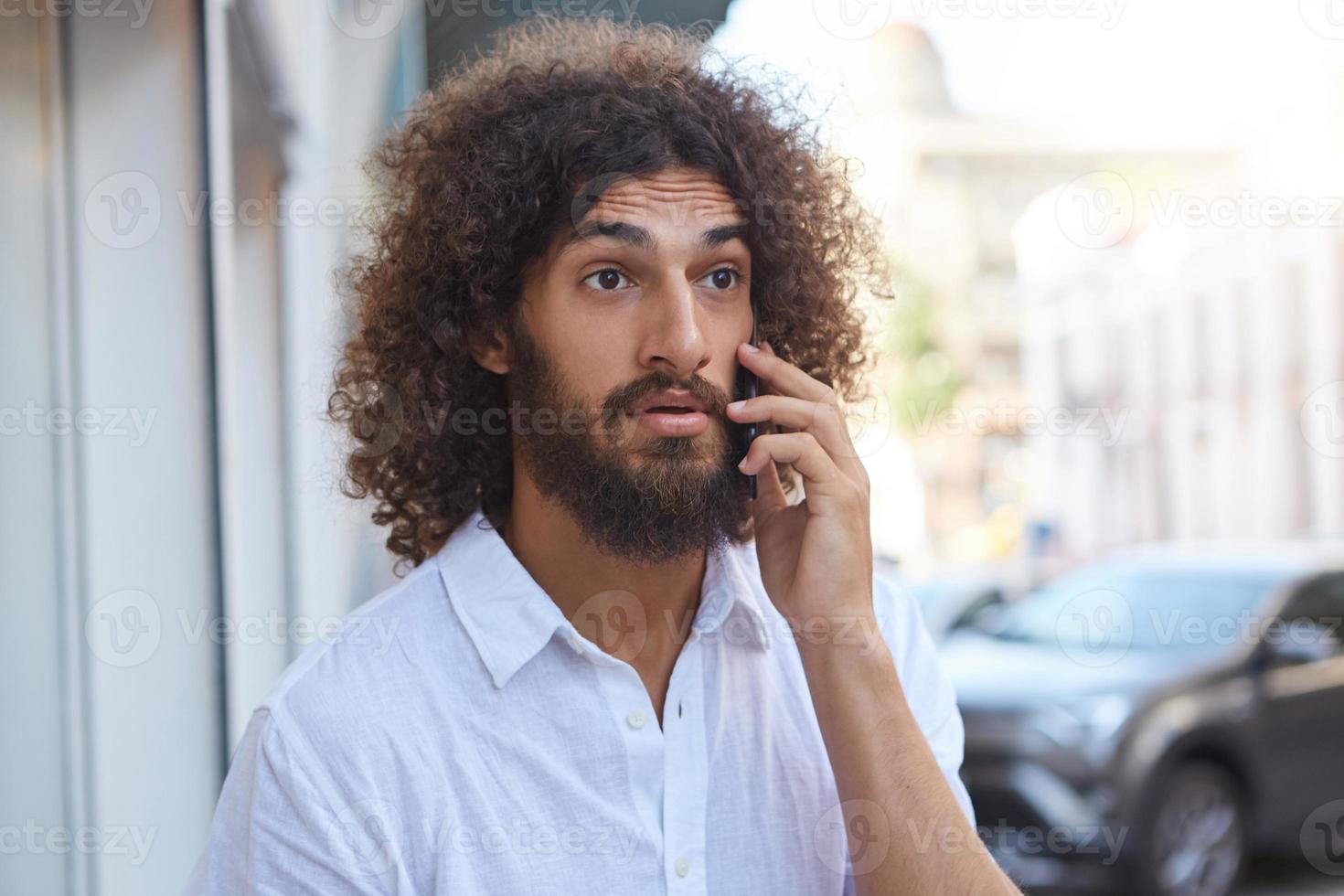 Surprised handsome bearded guy with brown curly hair walking down the street with mobile phone in hand, wrinkling forehead and raising eyebrows photo