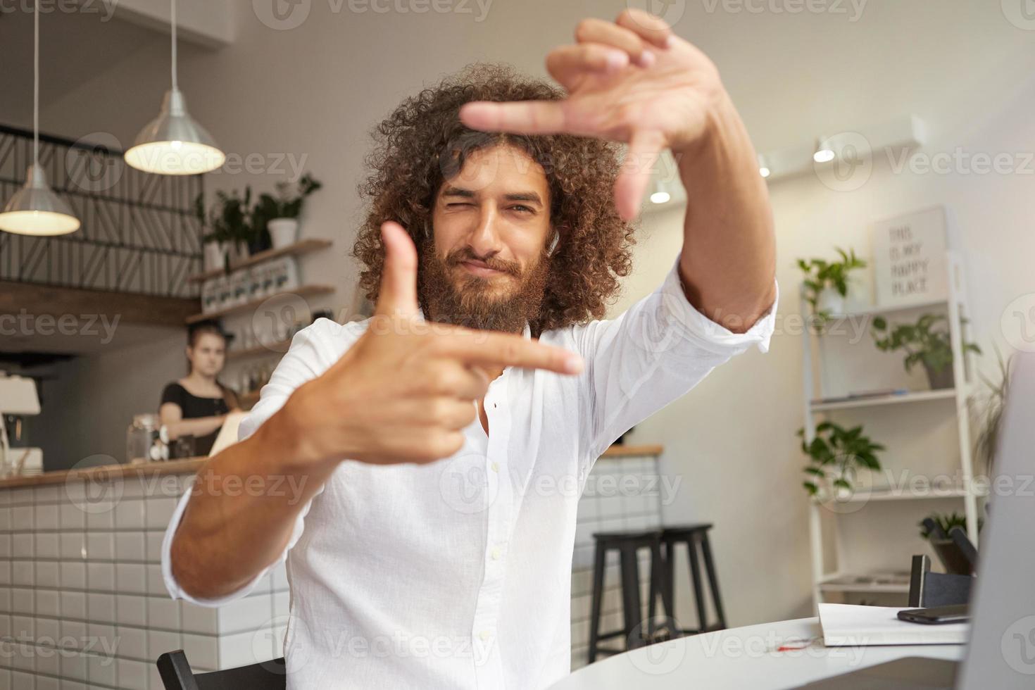 Young handsome bearded man with curly brown hair looking at camera and smiling, making frame gesture with hands, focusing using fingers with closed eye photo