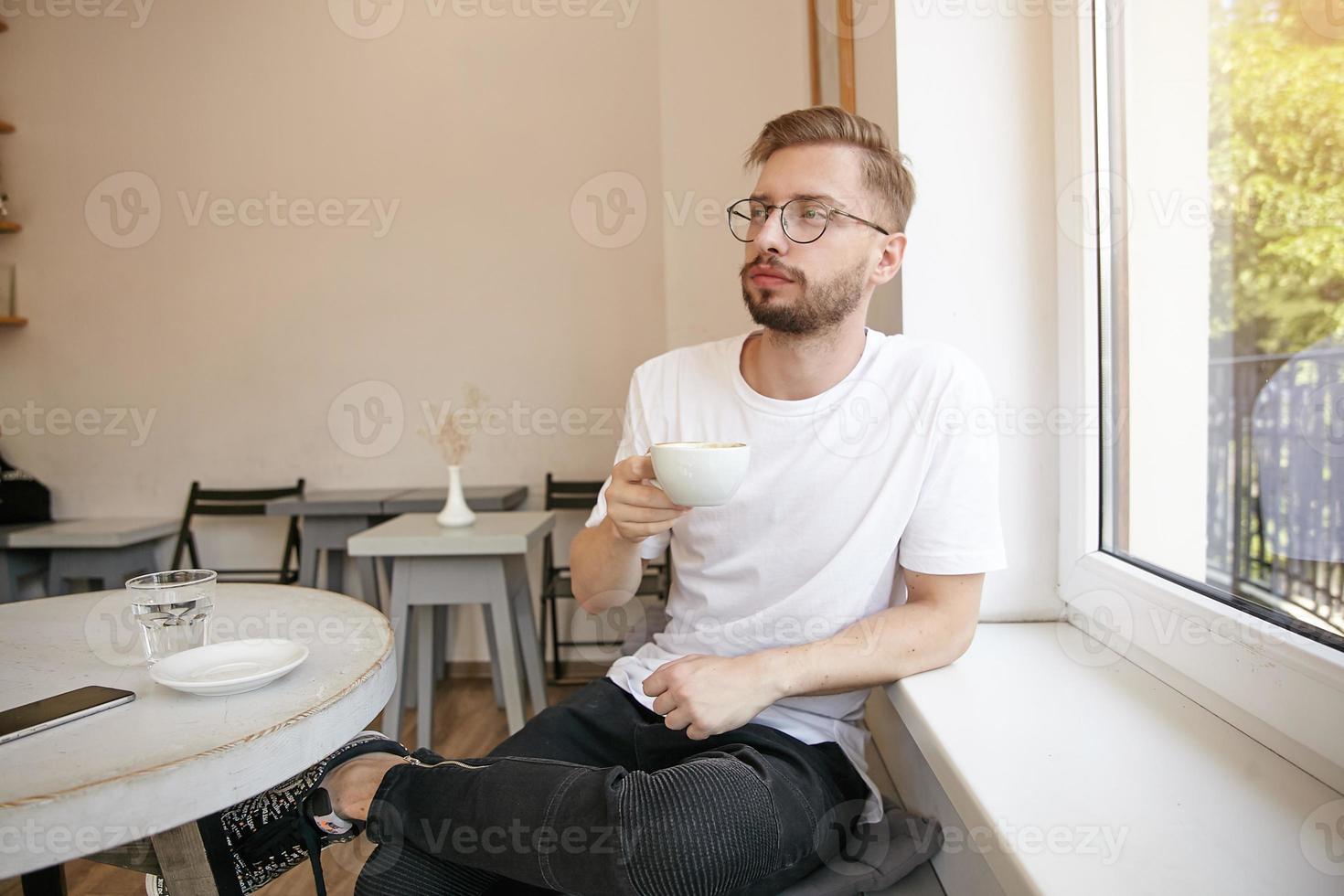 Portrait of young handsome male with beard, wearing casual clothes and glasses, sitting at the table in cafe with cup of coffee, waiting for somebody and looking thoughtfully photo