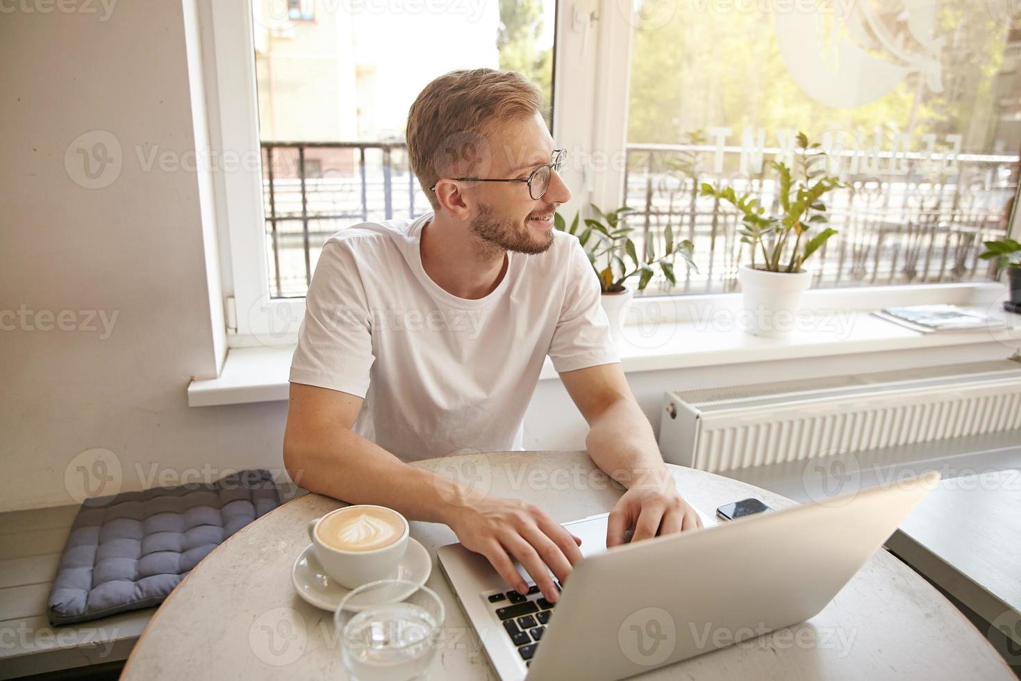 Interior portrait of good looking young bearded male is sitting at table with cup of coffee while working on laptop, smiling and looking dreamy aside photo