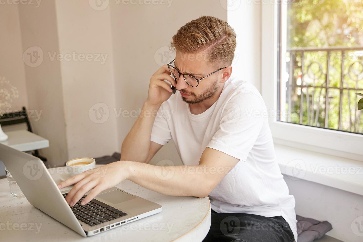 Portrait of handsome guy with short haircut solving problems on the phone, working remotely at public place, frowning and looking puzzled photo