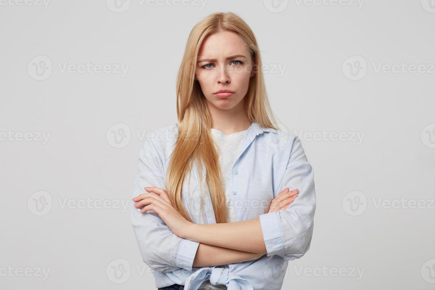 infeliz mujer rubia atractiva de pie sobre fondo blanco con los brazos cruzados, luciendo triste y a punto de llorar, usando ropa informal foto