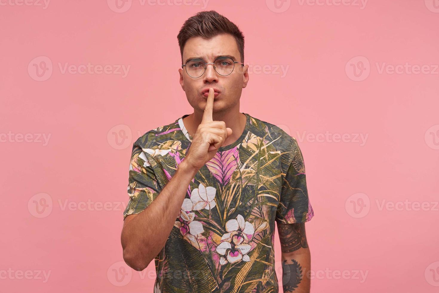 Serious good looking male with glasses keeping forefinger on lips, trying to keep conspiracy, calling to make silence, isolated shot over pink background photo