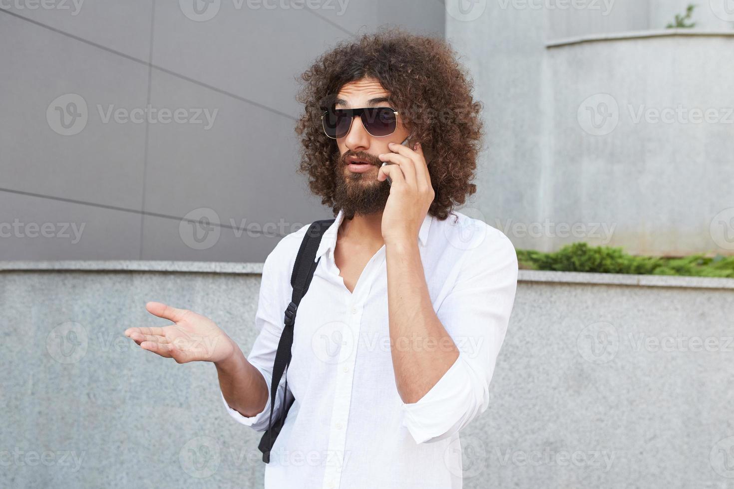 Wonders trucked young bearded man standing over outdoor background, raising his palm and wrinkling forehead, wearing glasses and white shirt photo