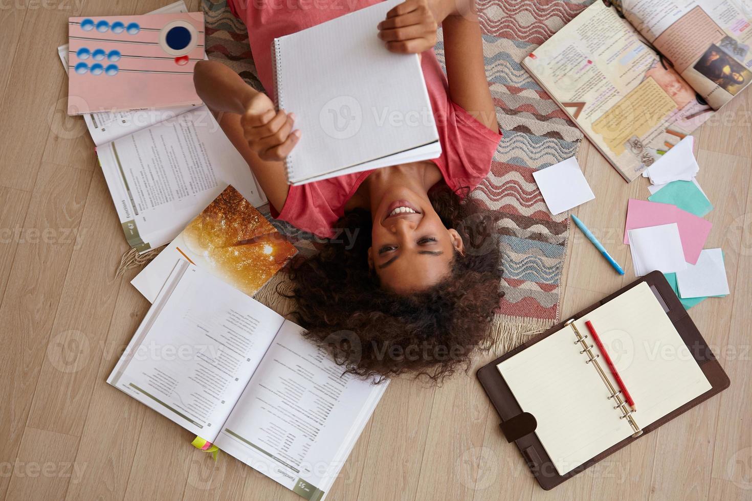 Top view of attractive young curly female with dark skin lying on carpet while reading notes, preparing for exams with a lot of books, wearing pink t-shirt photo