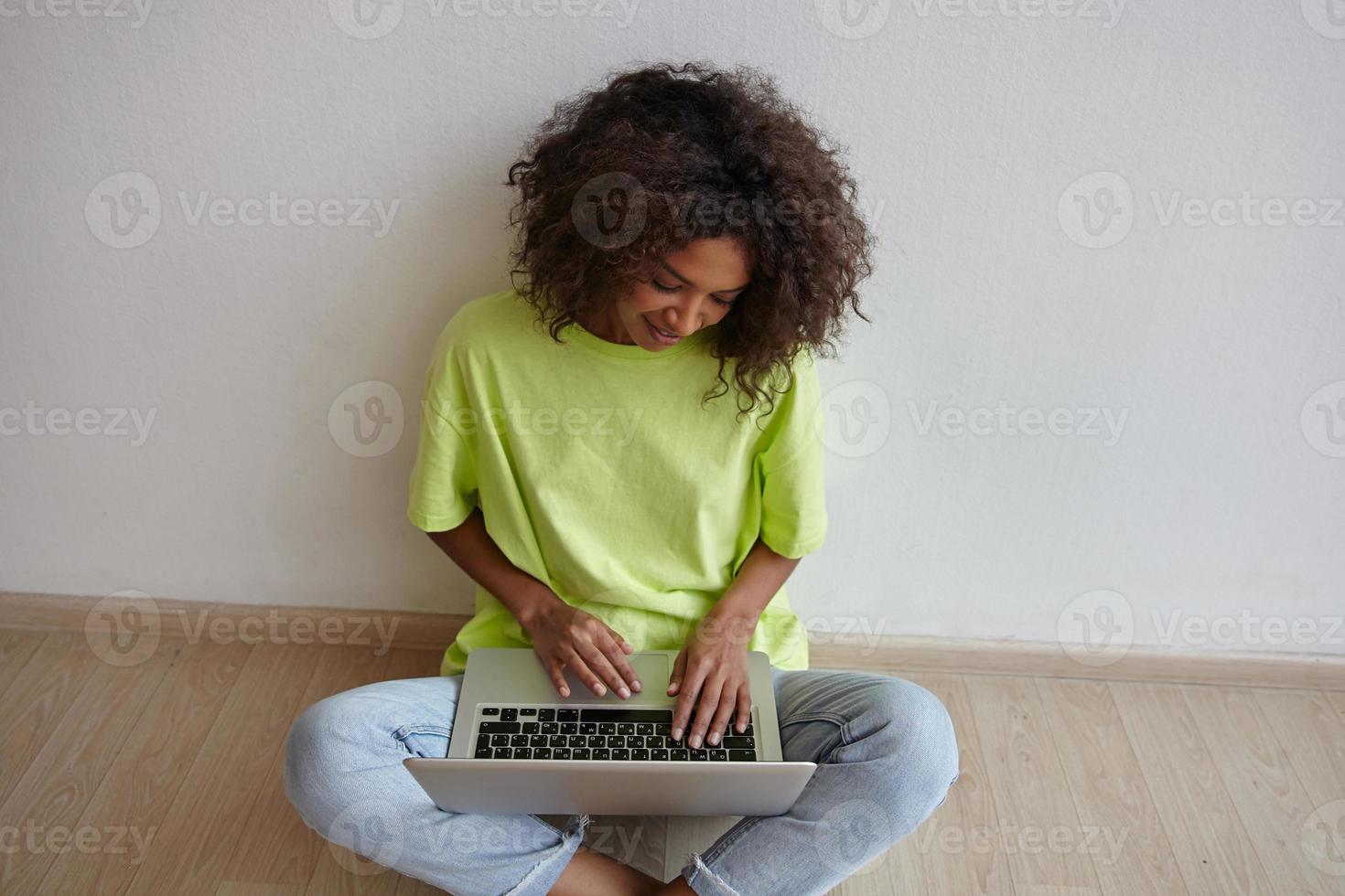 mujer joven y bonita independiente con cabello castaño rizado sentada en el suelo con las piernas cruzadas, manteniendo la computadora portátil en las piernas y trabajando en casa con ropa informal foto