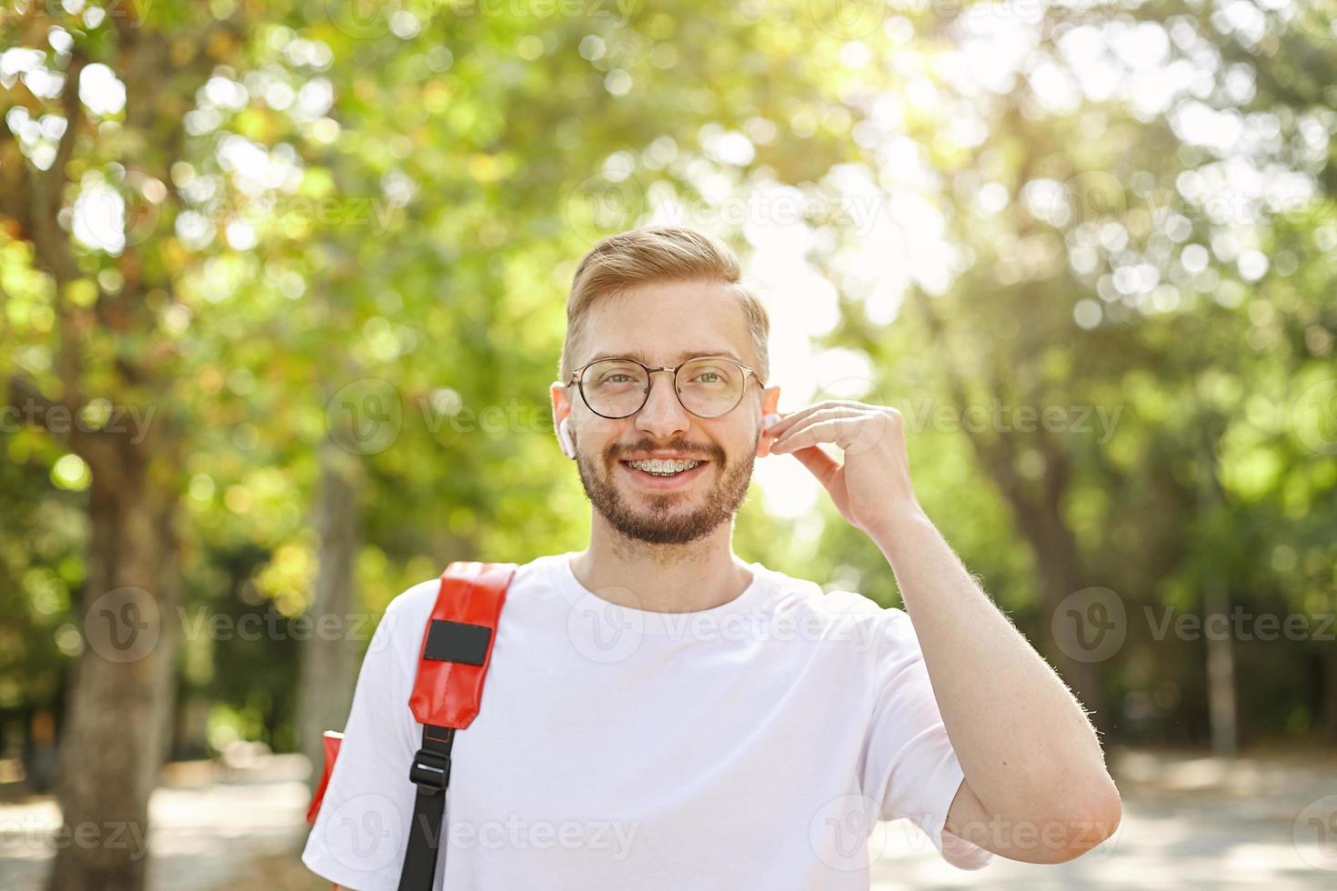 apuesto joven barbudo mirando a la cámara con una amplia sonrisa, usando anteojos y auriculares, siendo positivo y alegre, parado sobre árboles verdes foto