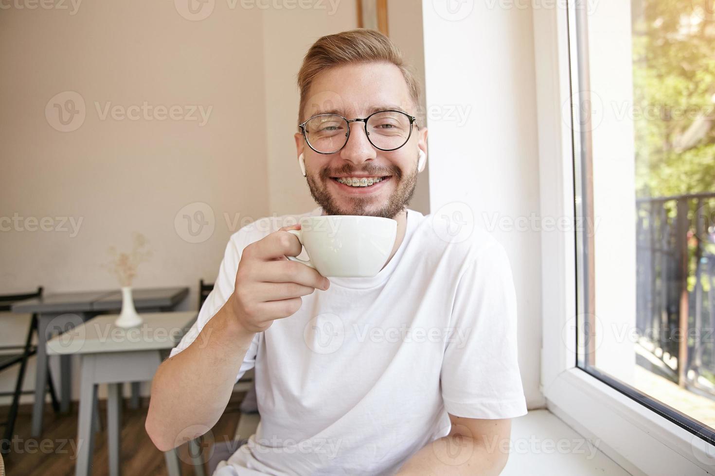 Indoor close-up of cute happy bearded guy, holding a cup in his hand and looking to camera with wide smile, wearing white t-shirt and glasses photo