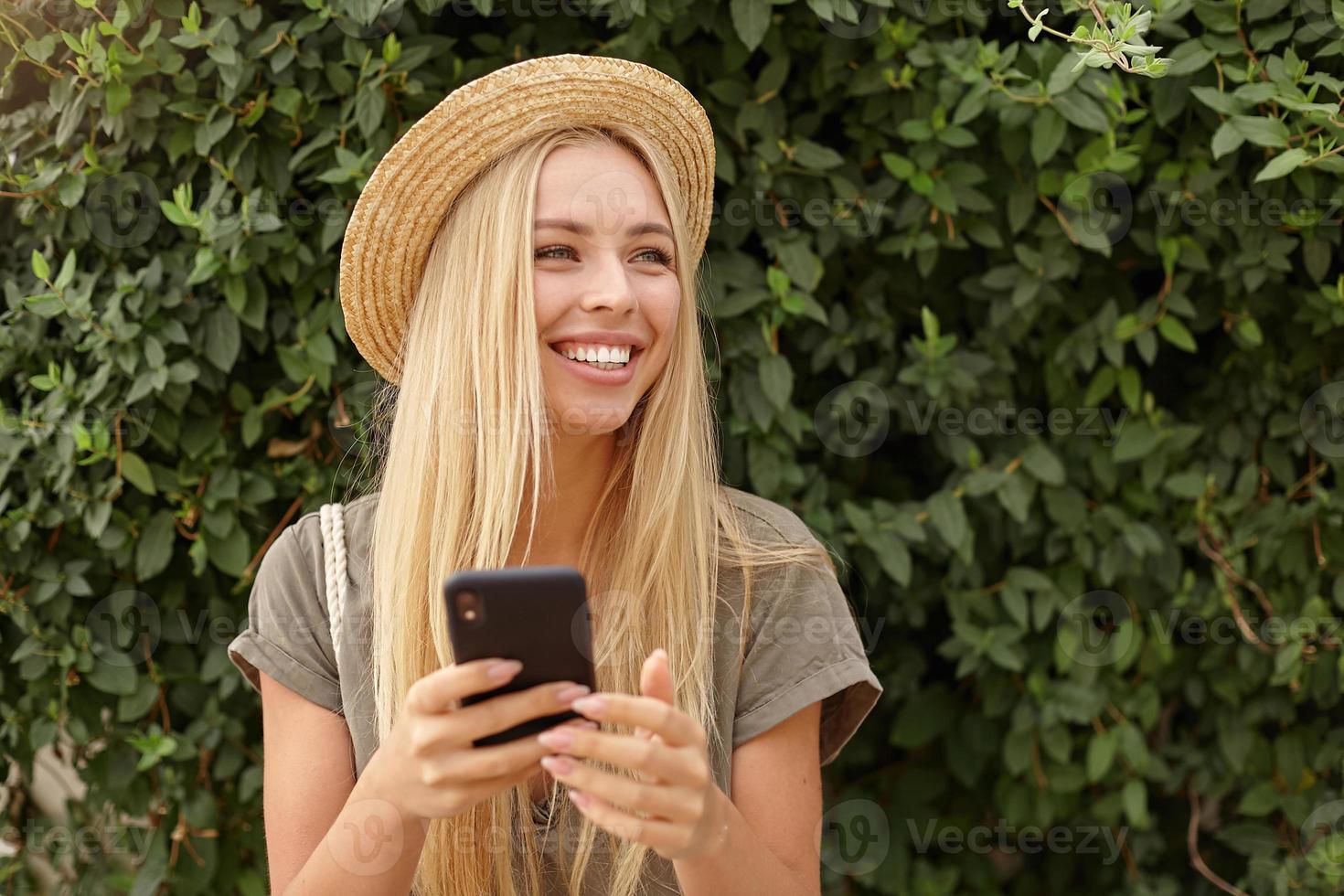 Smiling young beautiful woman looking aside happily, wearing casual clothes and straw hat, posing over green garden, keeping smart phone in hands photo