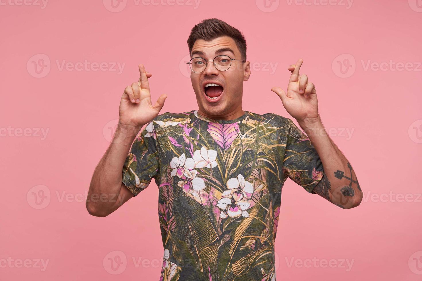 pretty young guy raising hands with crossed fingers for good luck, being excited about something, looking to camera with raised eyebrows and wide opened mouth, isolated over pink background photo