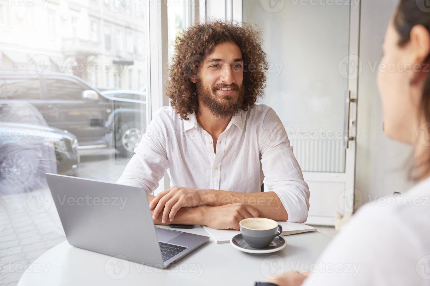 Cheerful young man with beard and brown curly hair meeting friend in coffee shop, working remotely with modern laptop, sitting at table near window with folded arms photo