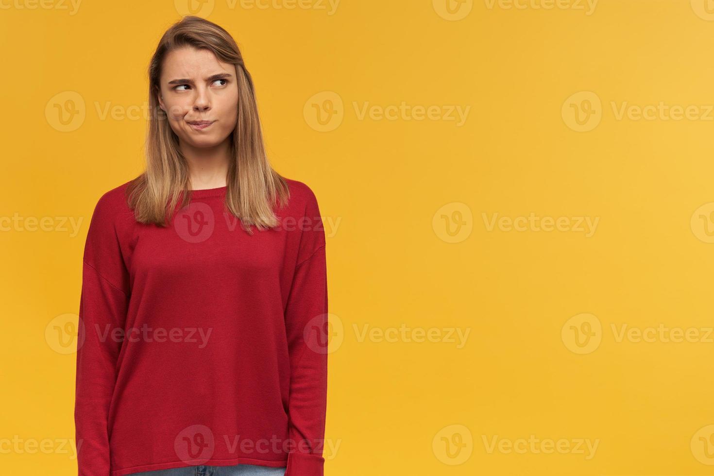Thoughtful girl with blond hair. Wearing red sweater. Lips pursed like doubting on some idea. Free copy space for your text. Looking to the upper right corner isolated over yellow background photo