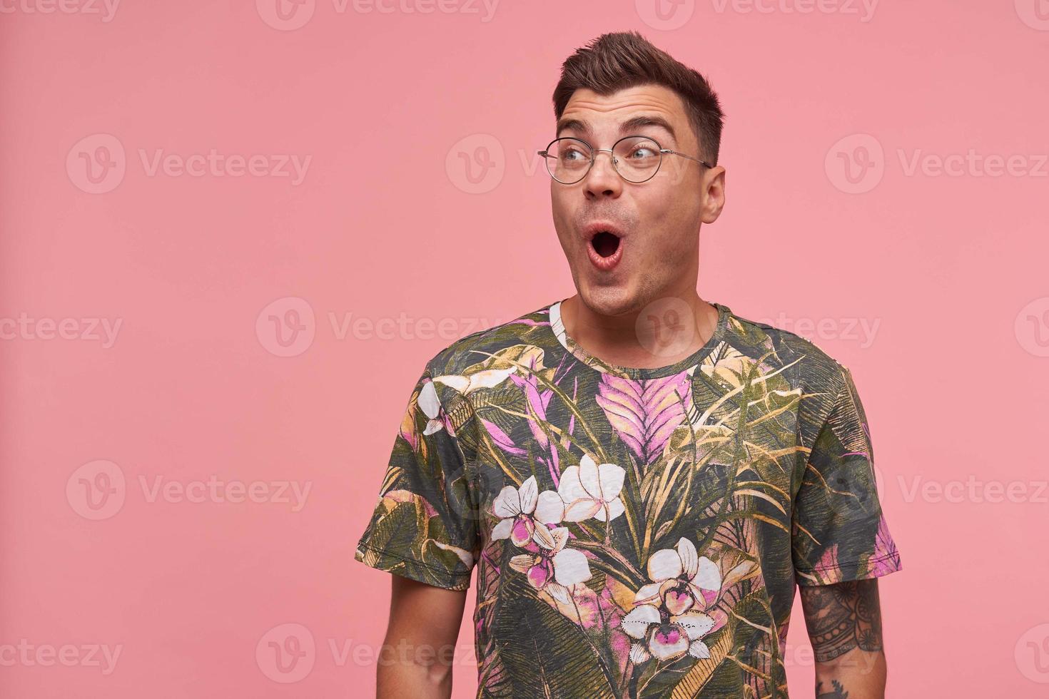 Studio portrait of handsome young lovely open-eyed male, looking aside with surprised face, puckering and folding lips, posing over pink background photo