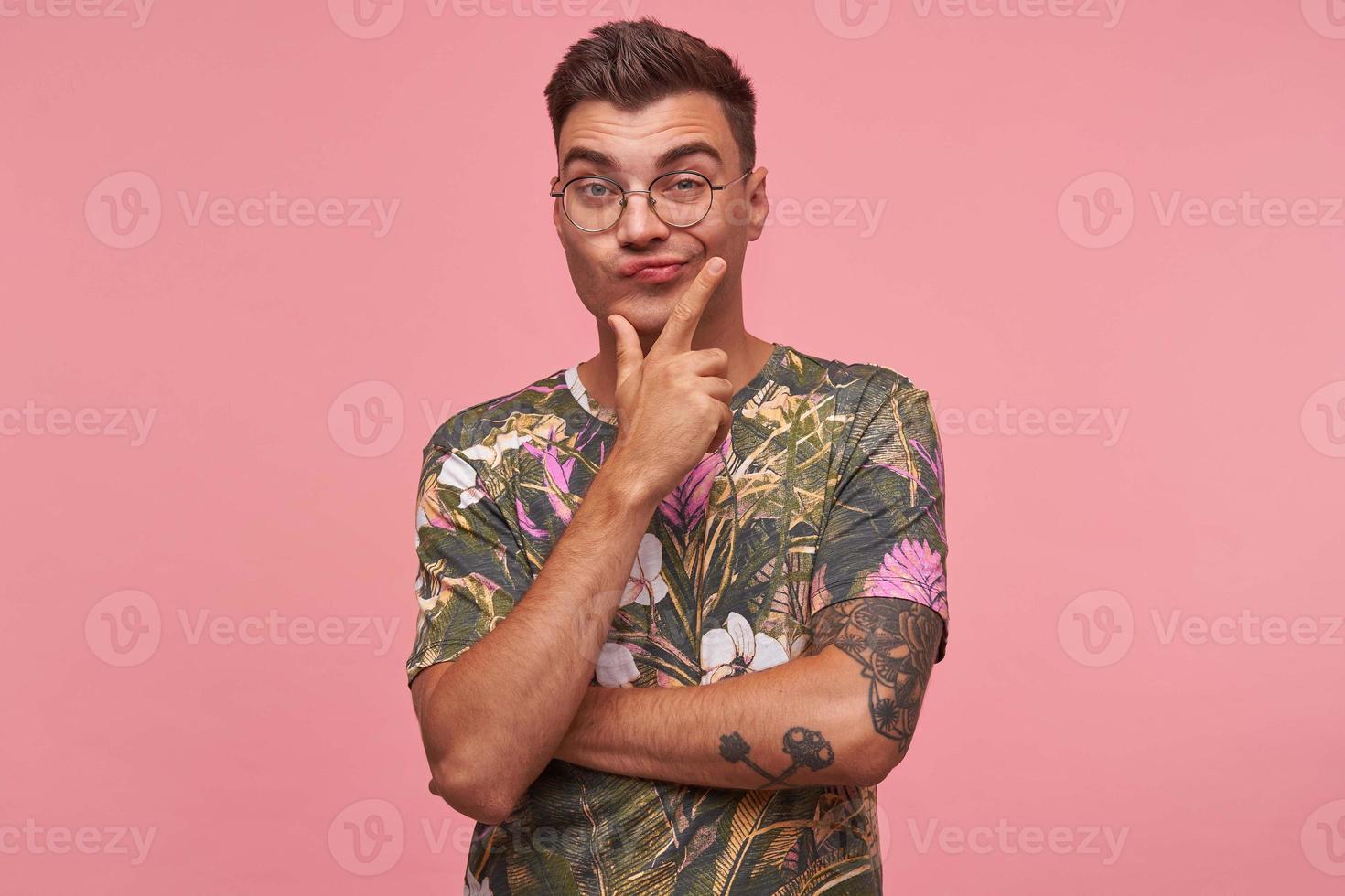 Indifferent handsome young dark haired male folding lips with raised eyebrows, holding his chin with apathetic face, posing over pink background photo