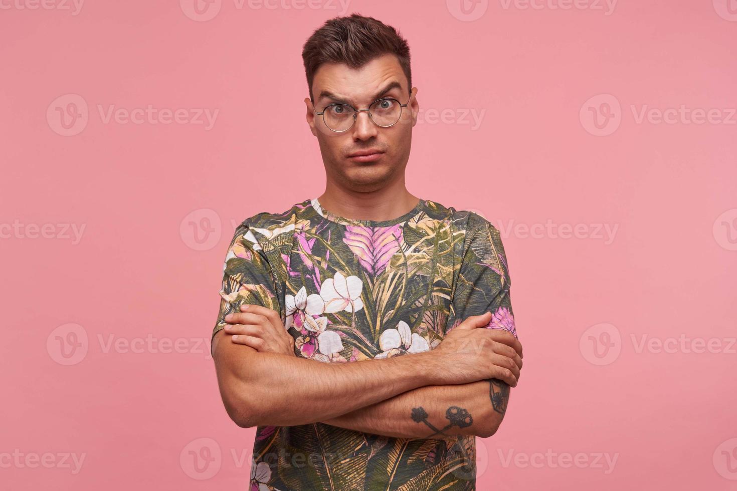 Indoor close-up of amazed pretty young male looking to camera with raised eyebrows, standing over pink background with crossed arms on his chest photo
