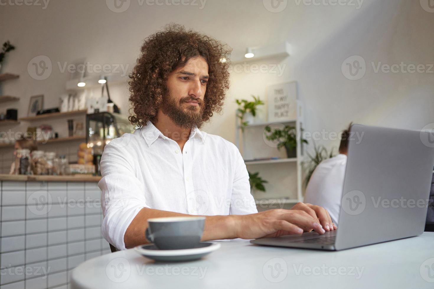 Indoor portrait of young serious businessman concentrating on work while sitting by desk in front of computer monitor, posing over cafe interior photo