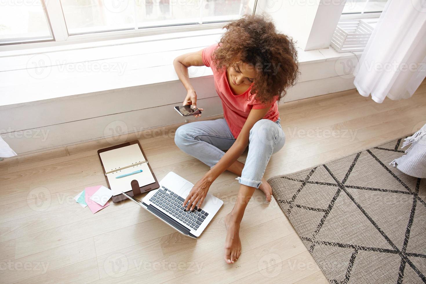 Top view of pretty dark skinned curly lady in casual clothes posing over home interior, working out of office with laptop and mobile phone, typing text on keyboard photo