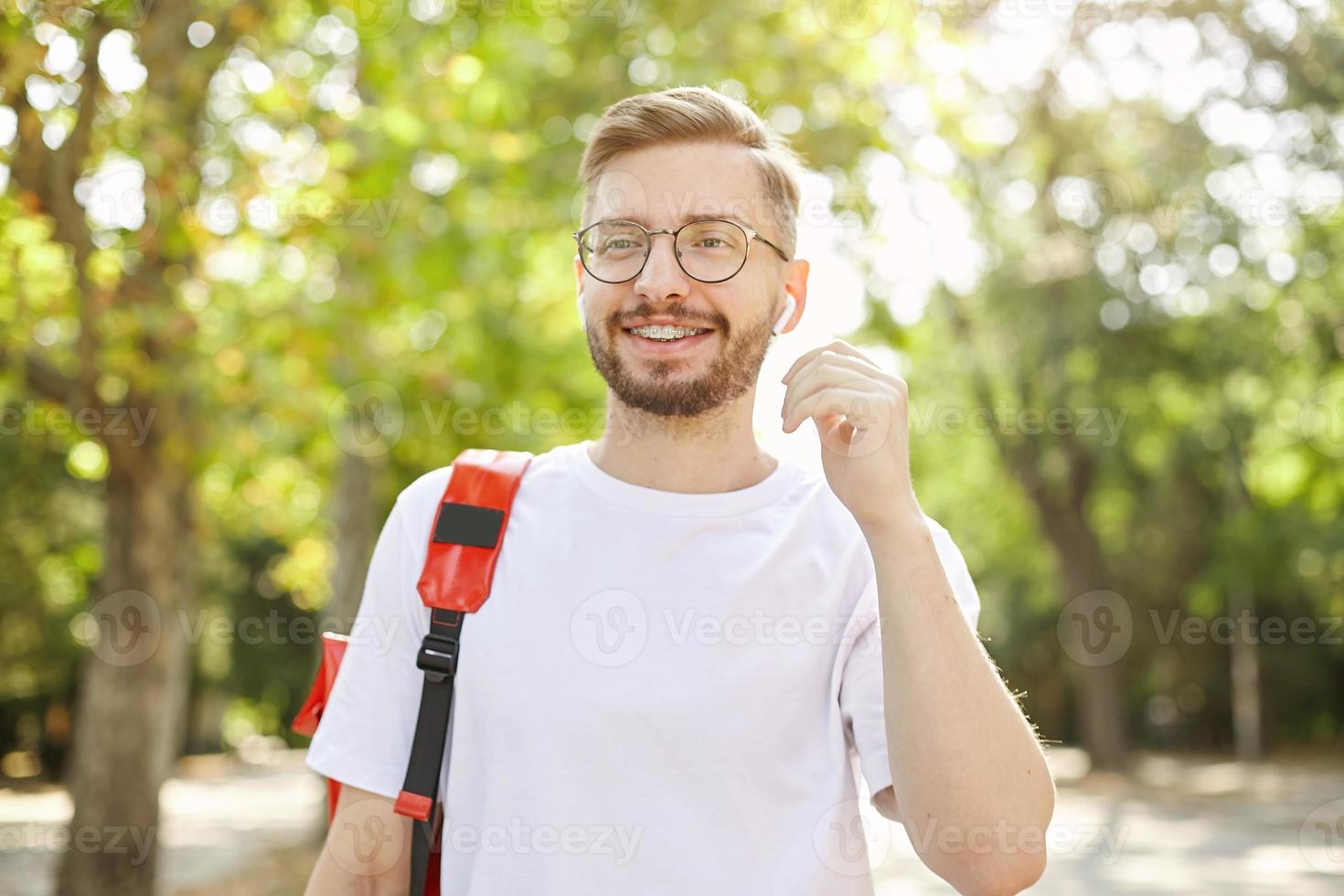 Outdoor shot of young handsome male with beard, wearing glasses and white t-shirt, smiling broadly and walking in park, going to pull out the earphone photo