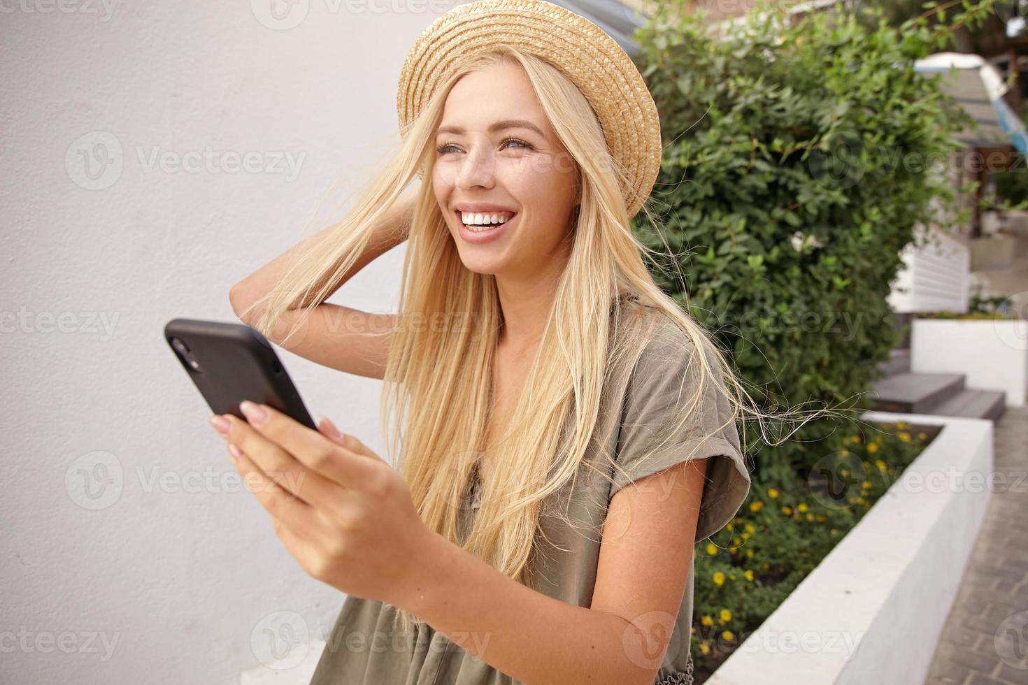 Outdoor shot of young charming woman with long blond hair straightening her straw hat, making selfie with smart phone, being happy and joyful photo