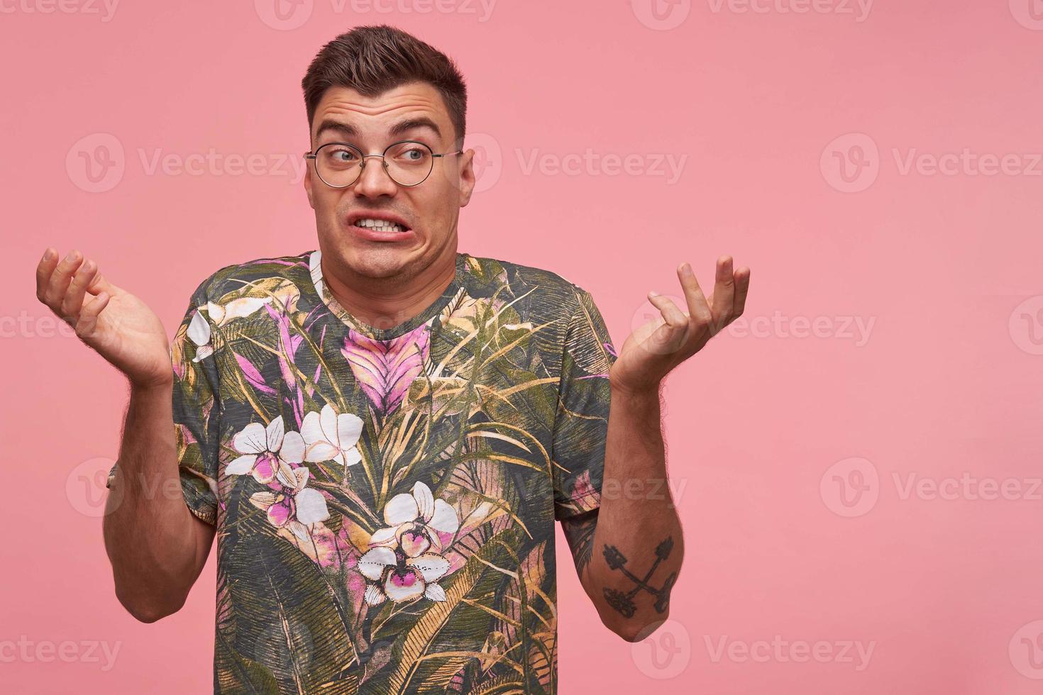 Portrait of handsome young male with eye wear looking aside with disgust, raising palms and making face, wearing t-shirt with floral print, isolated over pink background photo