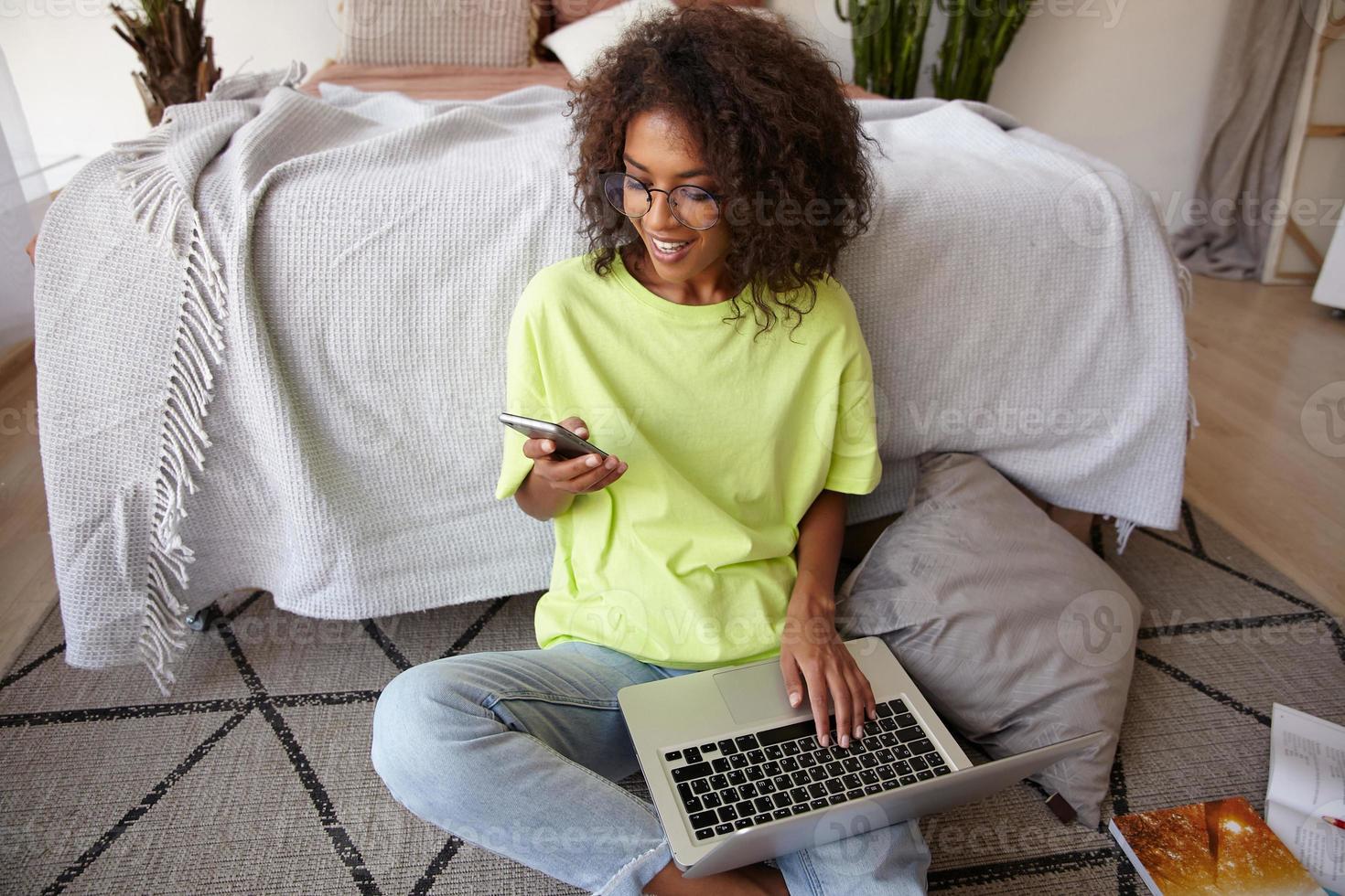 mujer joven de cabello oscuro de aspecto agradable con rizos trabajando en el piso del dormitorio, sosteniendo un teléfono inteligente en la mano y manteniendo la computadora portátil en las piernas, recibiendo buenas noticias, sonriendo alegremente foto