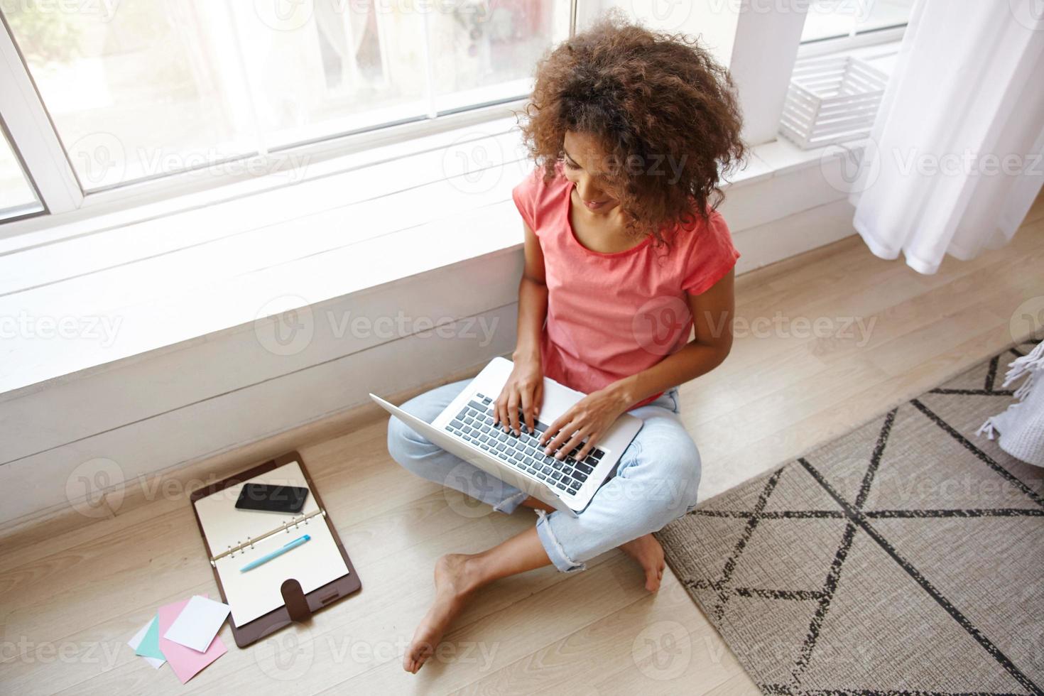 Indoor shot of young dark skinned female with brown curly hair sitting on floor with crossed legs, writing e-mail with her modern laptop, wearing casual clothes photo