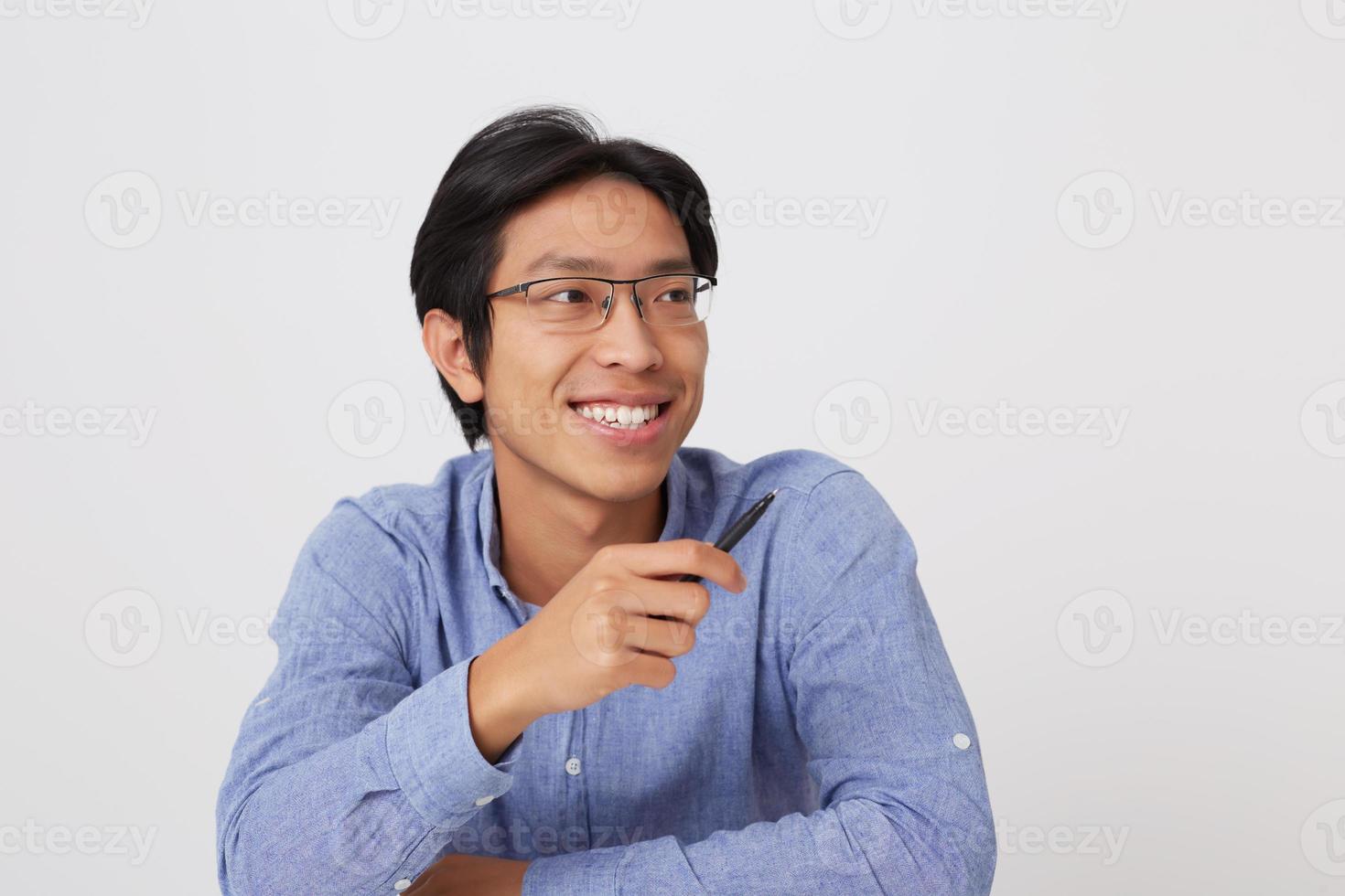 Portrait of happy handsome asian young business man in glasses and blue shirt pointing to the side by pen isolated over white background photo