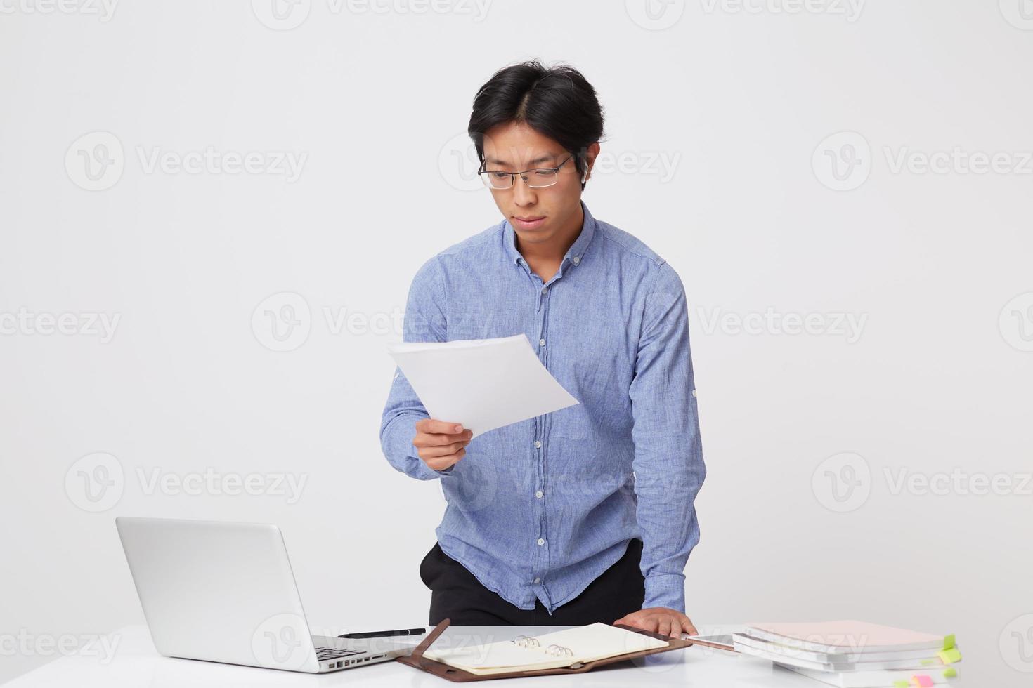 Concentrated asian young business man in glasses using laptop and earphones for video communication working with documents over white background photo