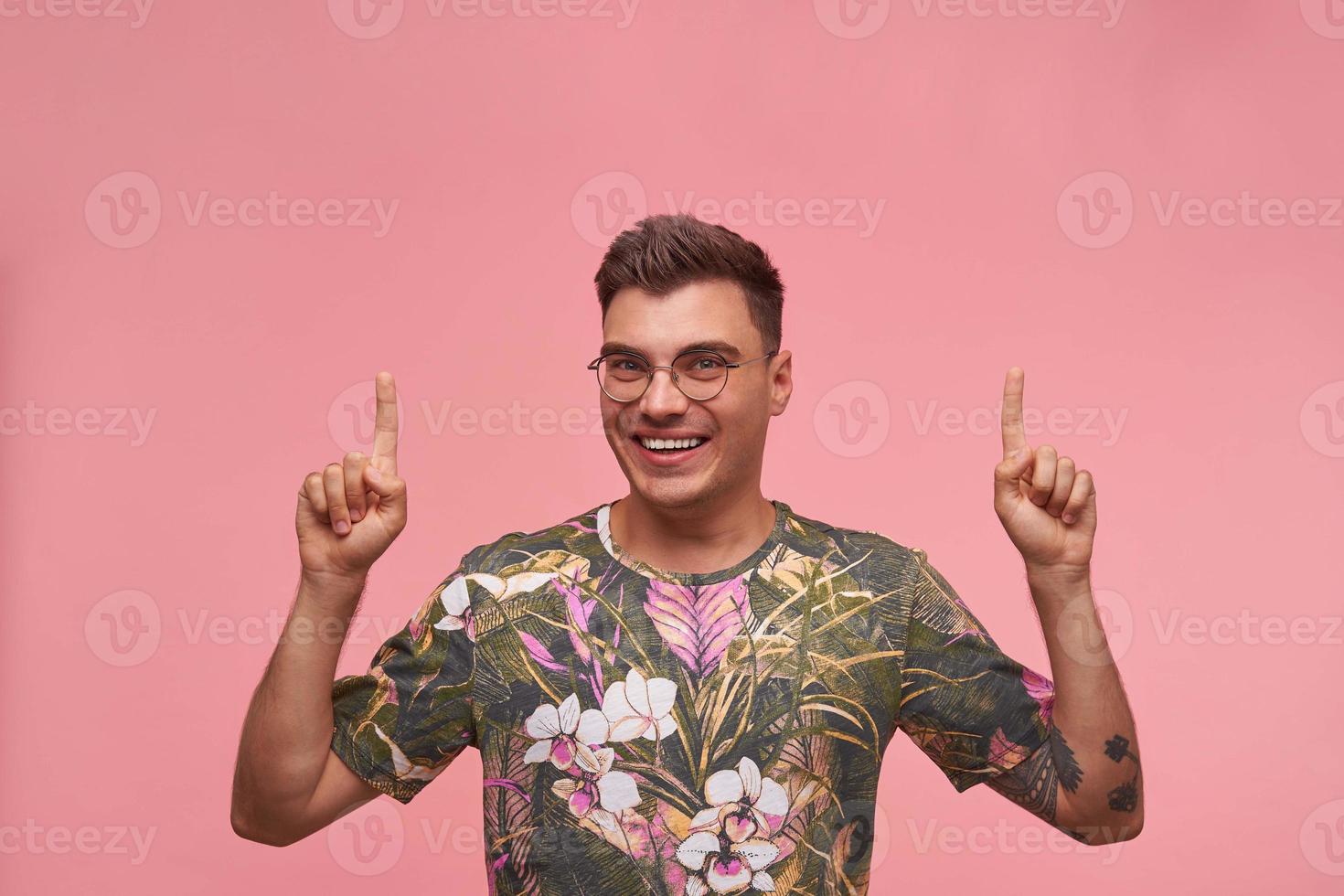 Happy young dark haired man in casual t-shirt smiling to camera with raised hands, pointing with fingers upwards, standing over pink background photo