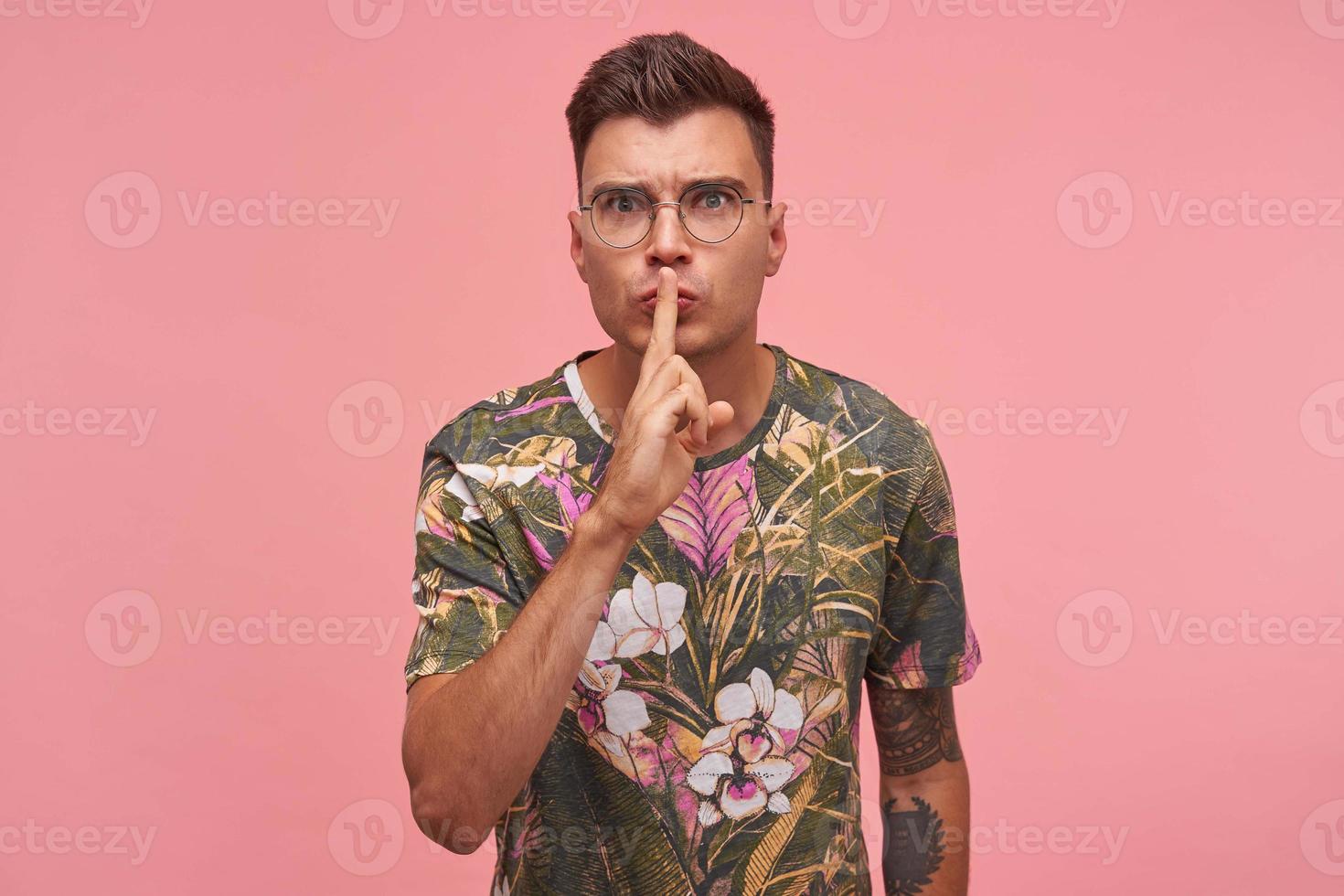 Close up of attractive young man with tattoos keeping index finger on lips, asking to keep secret, do not make noise, demonstrating silence gesture, standing over pink background photo