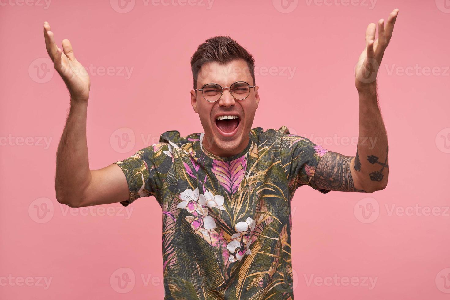 Indoor portrait of agitated pretty short haired young man looking to camera with wide mouth opened, being excited and nervous, raising hands up and shouting, isolated over pink background photo