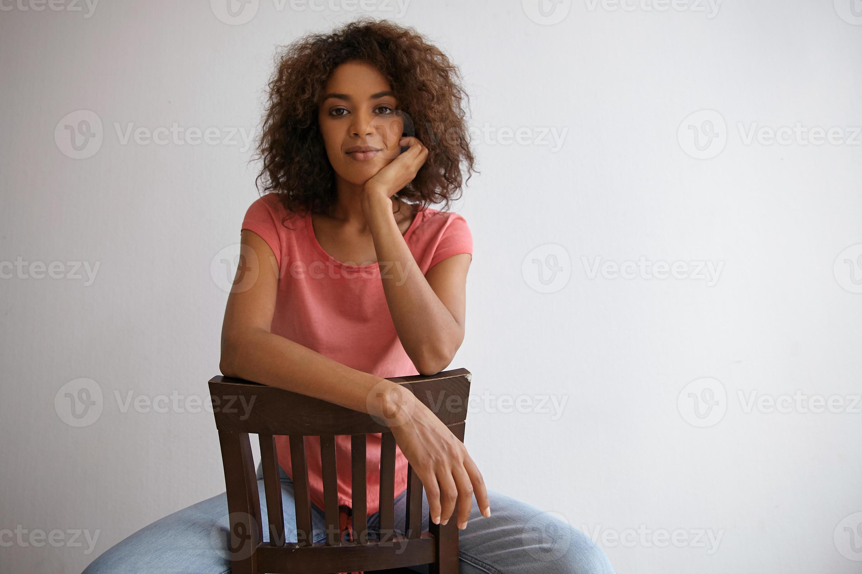 Good looking young dark haired curly female posing over white background in  casual clothes, sitting in front of camera with hand on back chair and  chinning up 9247991 Stock Photo at Vecteezy