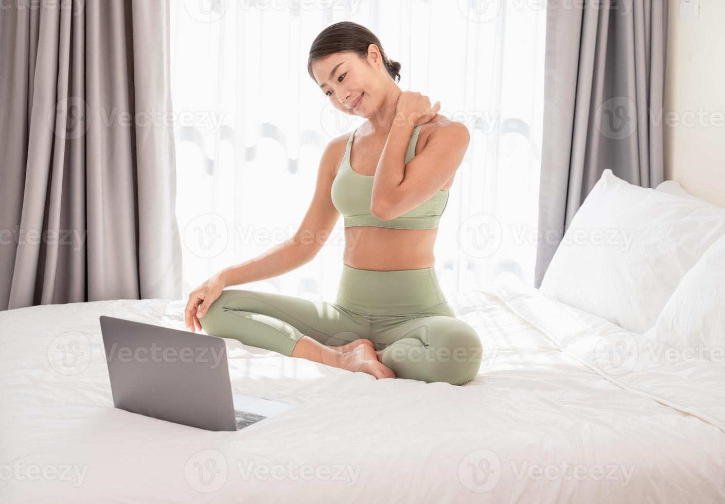 Woman practicing yoga on bed after waking up. Asian female sitting on bed using her laptop while doing stretching exercise at home. photo