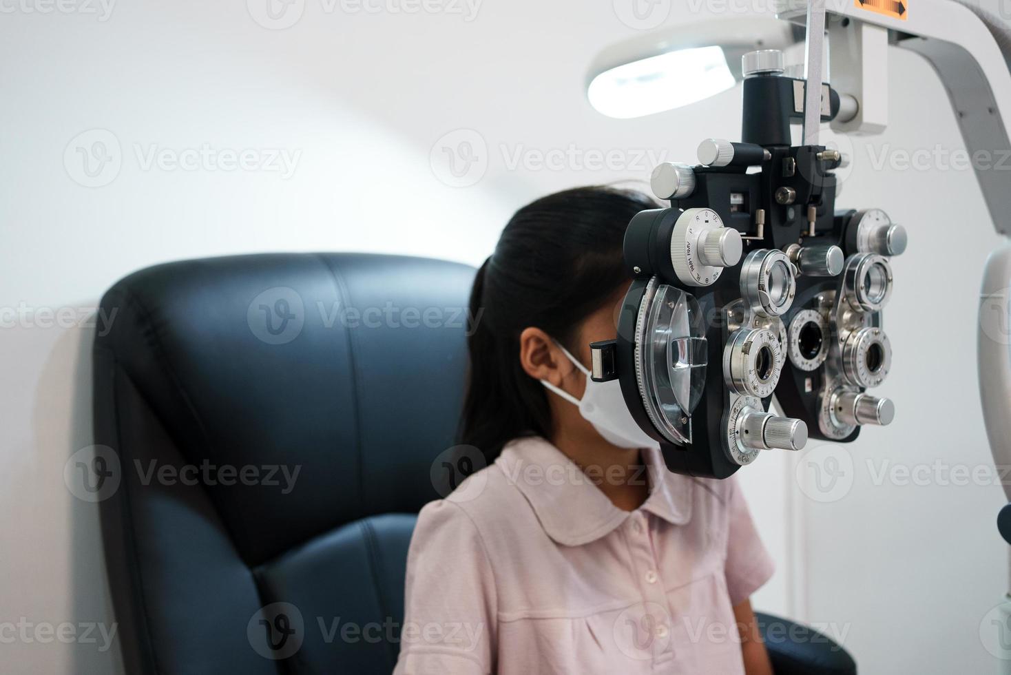 Ophthalmologist examining the eyes of an Asian girl in a clinic. They wear protective face masks. photo