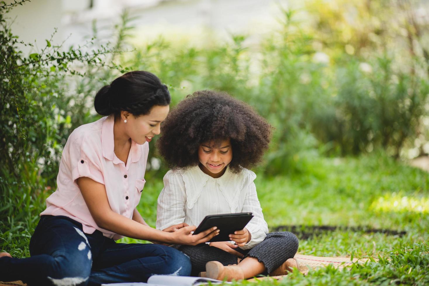 Cheerful young beautiful mother sitting with son with curly hair in park and helping him while using digital tablet while studying photo