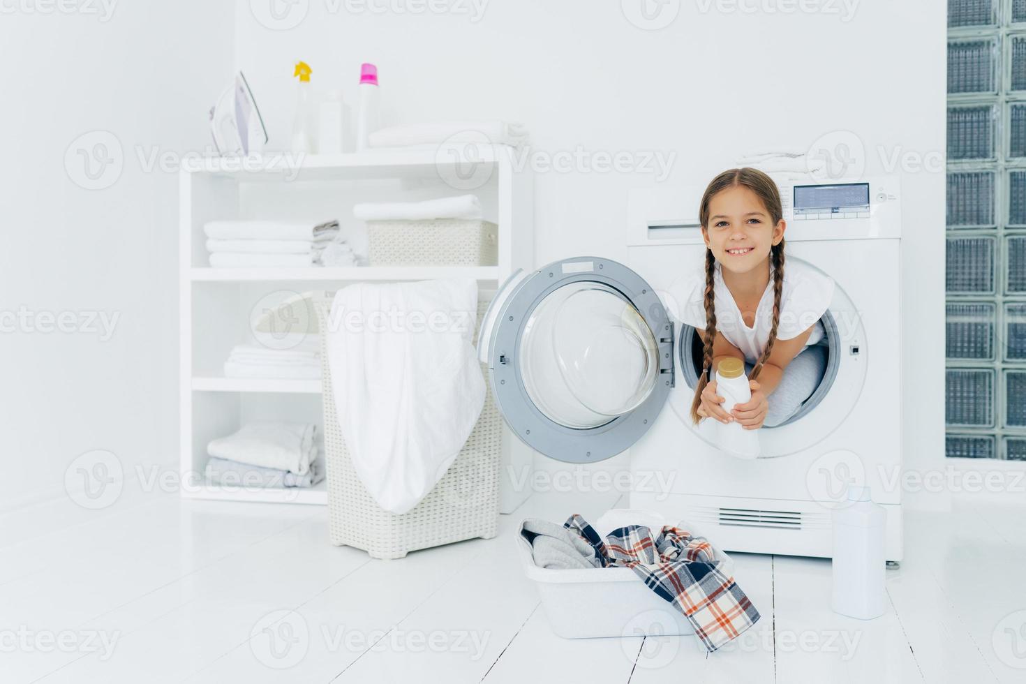 niña pequeña con apariencia atractiva, se divierte y posa dentro de la lavadora, sostiene detergente, se prepara para lavar, lavabo con ropa para poner en lavadora en piso blanco. día de lavado en casa foto