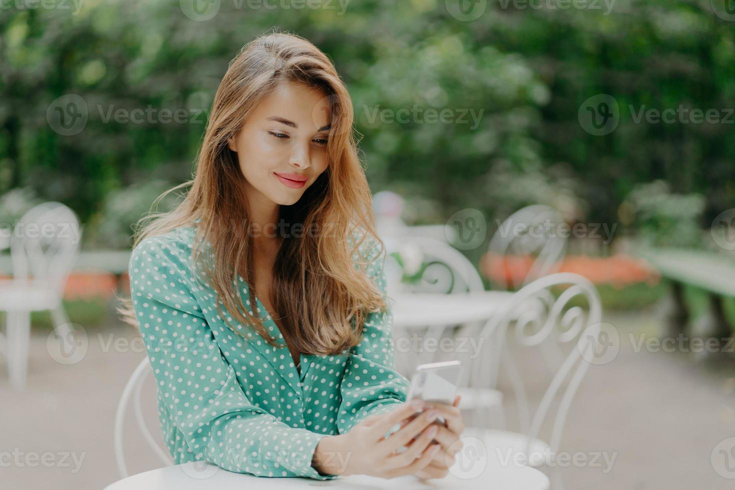 hermosa mujer con maquillaje, vestida con ropa de moda, vestida con un pantalones de lunares de moda, usa el teléfono móvil para comunicarse en línea, navega por las redes sociales, posa en una cafetería al aire libre foto