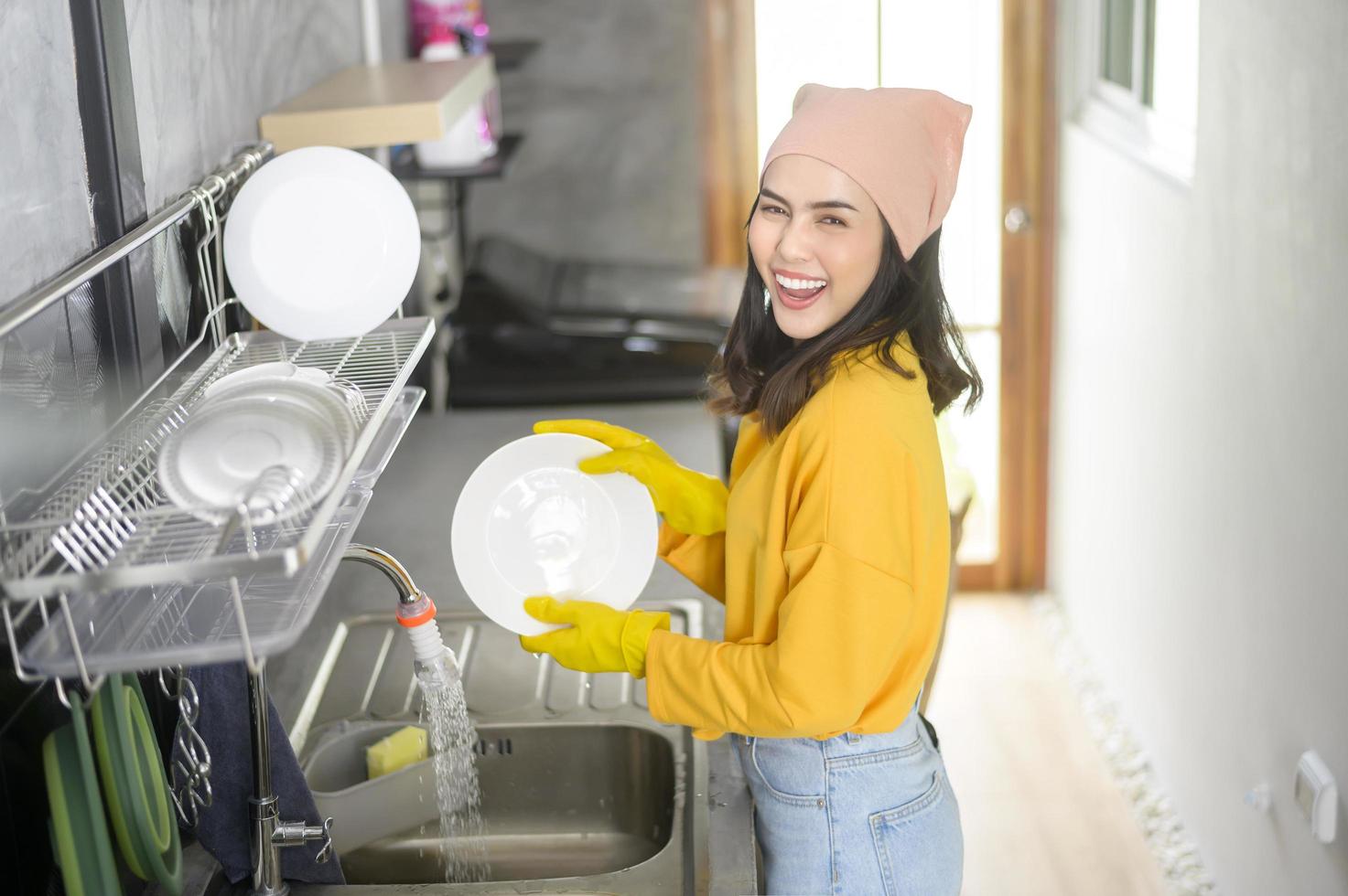 joven mujer feliz usando guantes amarillos lavando platos en la cocina en casa foto