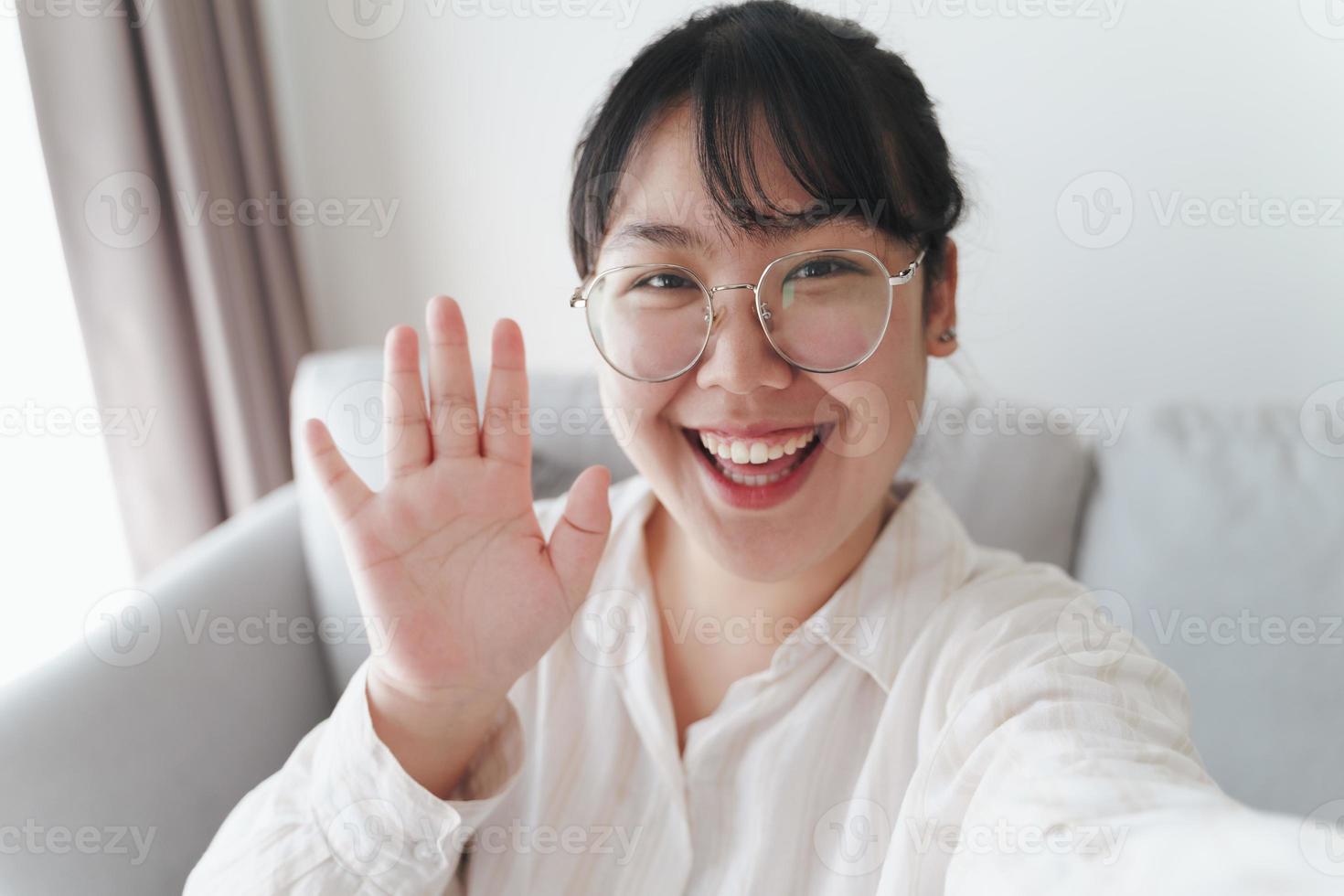 Young Asian woman using smartphone for online video conference call with friends waving hand making hello gesture photo