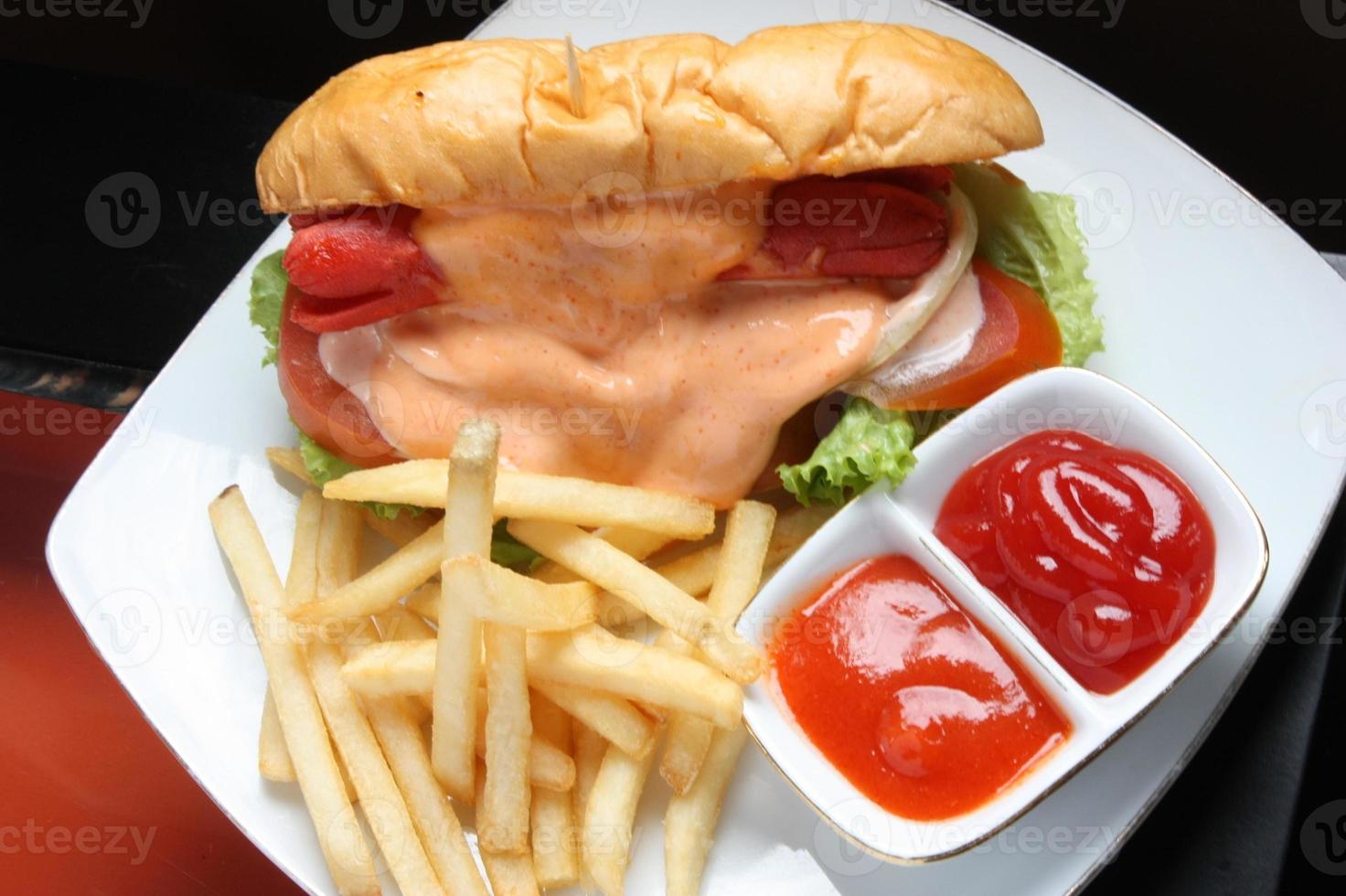 Complete hotdog package. Red sausage, cheese sauce, tomato wedges, lettuce. Plus french fries and a container of red chili and tomato sauce. Served on a white square plate. Close up. top view photo