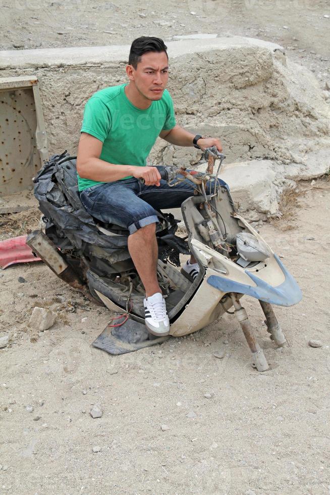 A young man with dark hair sits on a wrecked scooter on a parking lot in Mykonos, Greece. photo