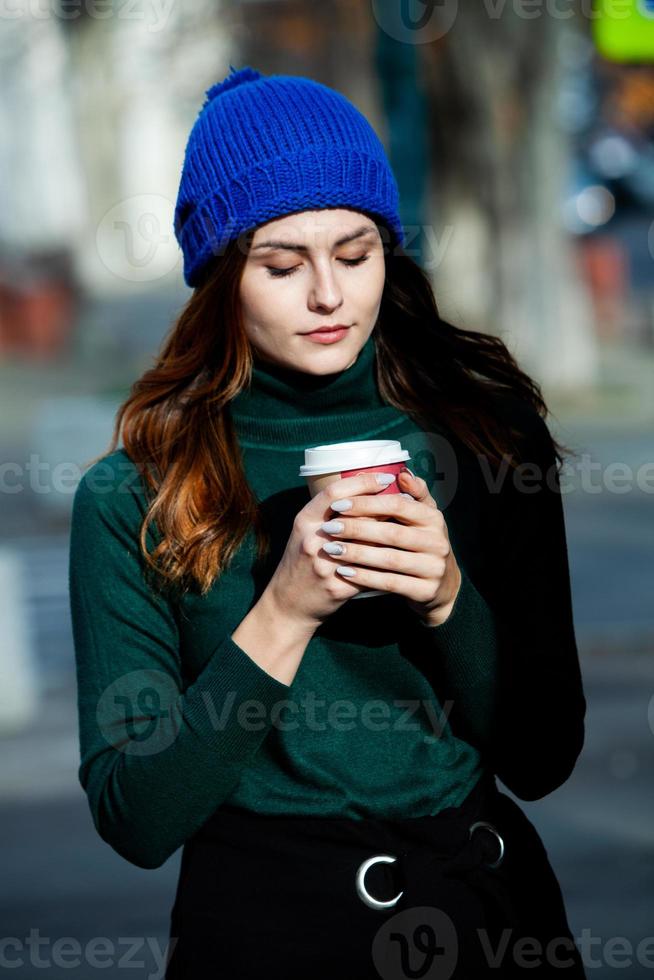 Young stylish woman drinking tea in a city street. Hipster european girl with latte paper glass. Gorgeous young woman with cup of coffee in city street. Coffee break. Coffee to go. photo