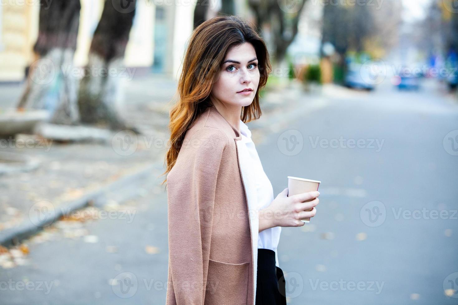 joven elegante bebiendo té en una calle de la ciudad. hipster chica europea con vaso de papel con leche. hermosa mujer joven con una taza de café en la calle de la ciudad. descanso. Café para llevar. foto