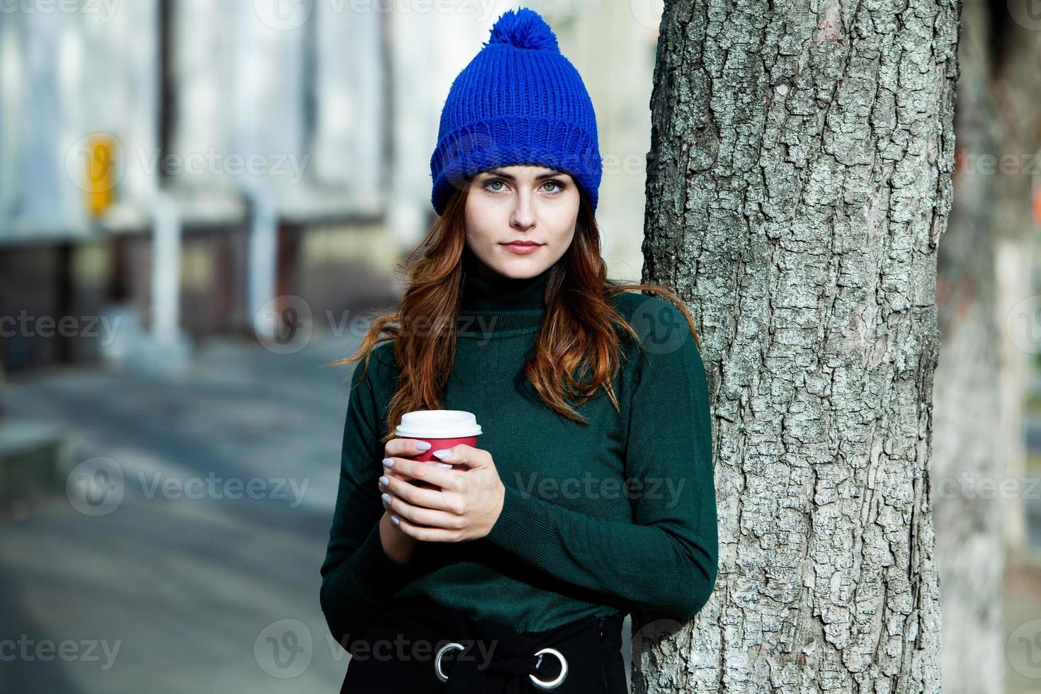 joven elegante bebiendo té en una calle de la ciudad. hipster chica europea con vaso de papel con leche. hermosa mujer joven con una taza de café en la calle de la ciudad. descanso. Café para llevar. foto