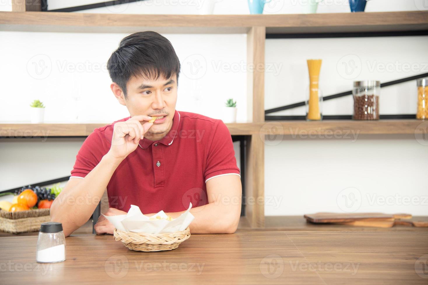 Asian syoung man with casual  red t-shirt enjoy having breakfast, eating french fries. Young man cooking food  in the loft style kitchen room photo
