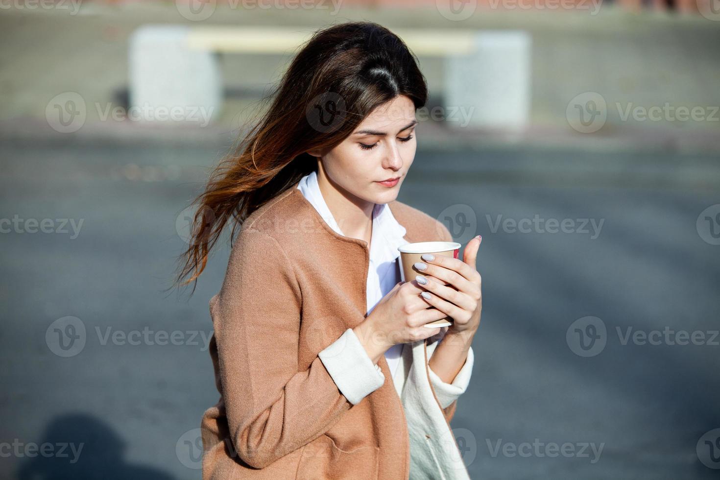 joven elegante bebiendo té en una calle de la ciudad. hipster chica europea con vaso de papel con leche. hermosa mujer joven con una taza de café en la calle de la ciudad. descanso. Café para llevar. foto