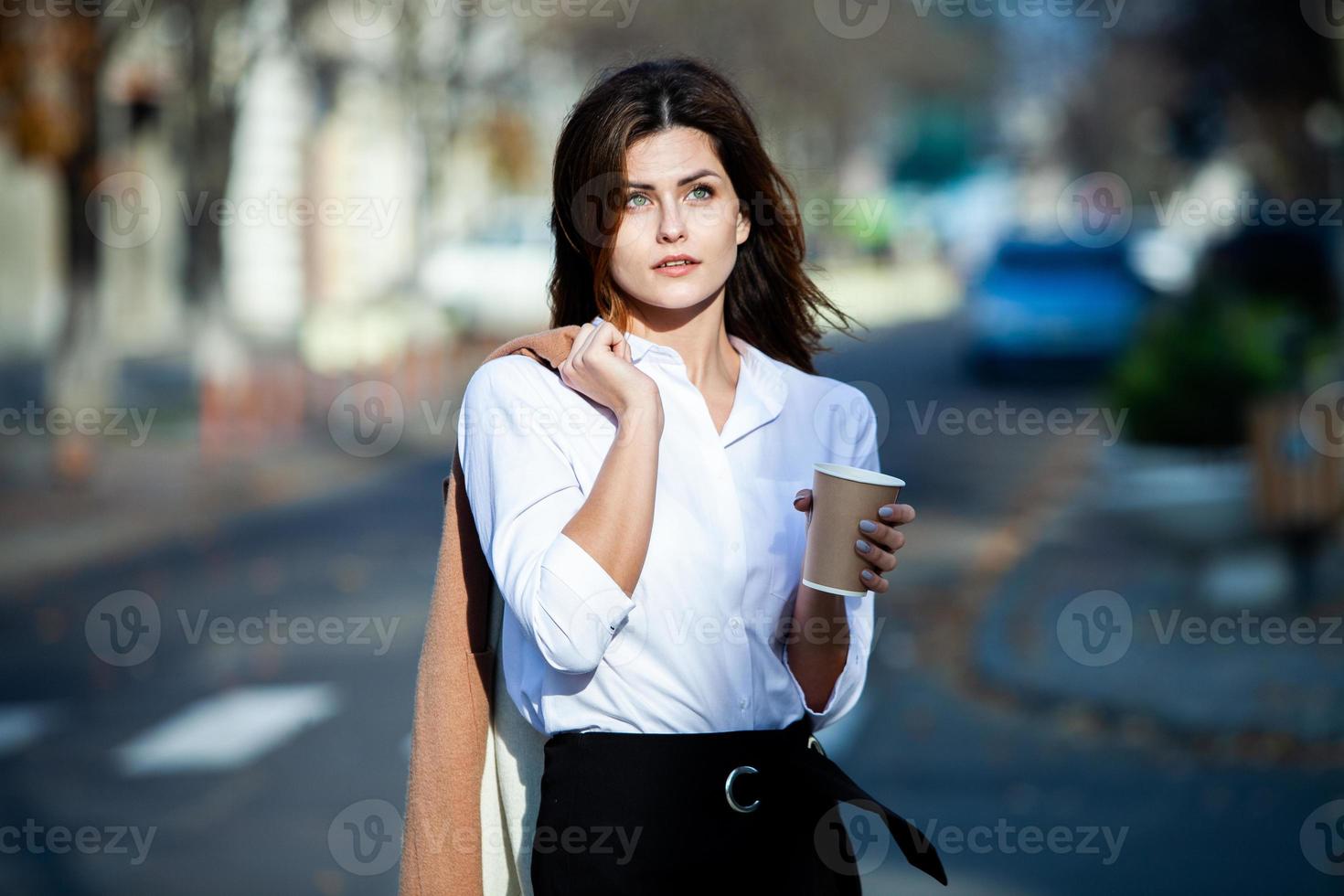 joven elegante bebiendo té en una calle de la ciudad. hipster chica europea con vaso de papel con leche. hermosa mujer joven con una taza de café en la calle de la ciudad. descanso. Café para llevar. foto