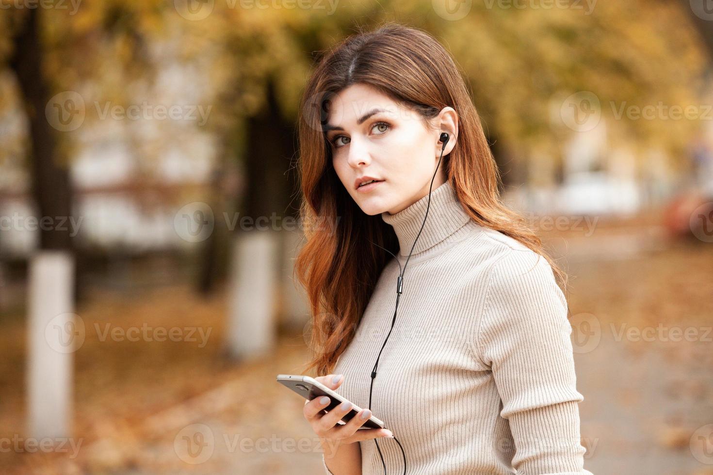 Image of beautiful stylish woman holding mobile phone. Young european girl standing at the street and using cellphone. Woman listening to music with the phone and having fun. Mobile internet concept. photo