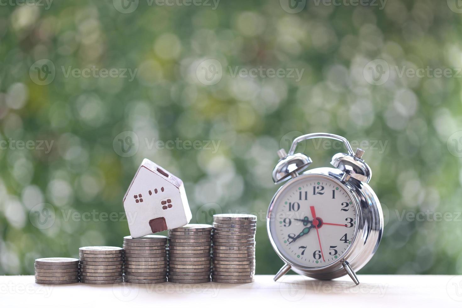 Model house on stack of coins money and alarm clock on natural green background, investment and business concept photo